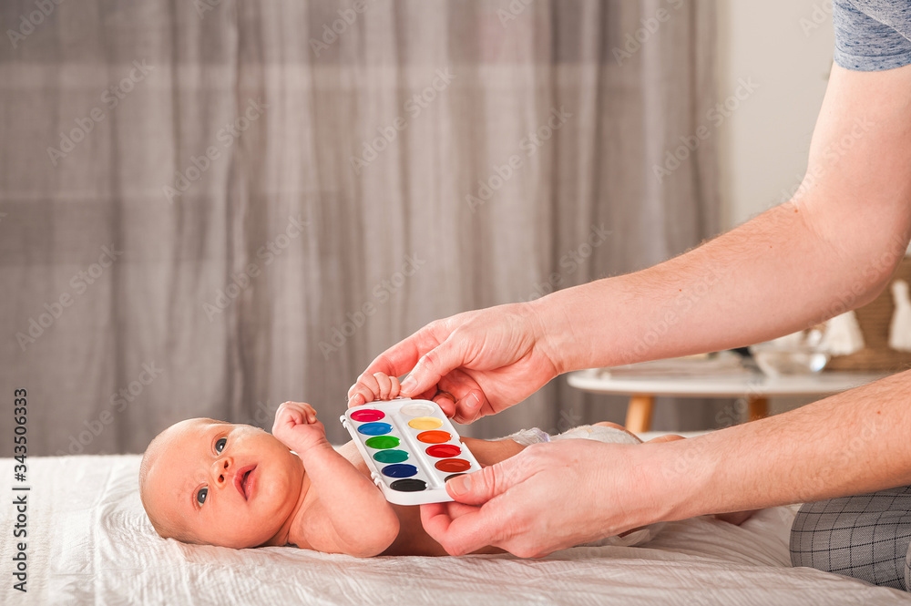 Naklejka premium Newborn with paints on a white plaid closeup. Infant holds watercolor rainbow paint and copy space. The concept of the game, care and care for the baby. 