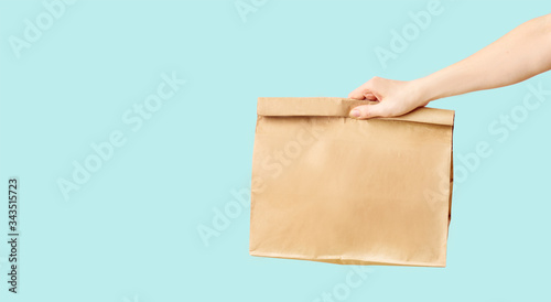 Female hand receiving brown clear empty blank paper bag for food delivery on blue background with copy space. Packaging template mock up fast food.