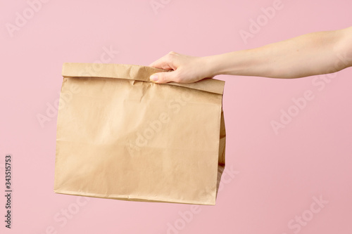 Female hand holding brown clear empty blank paper bag for food delivery on pink background with copy space. Packaging template mock up fast food.