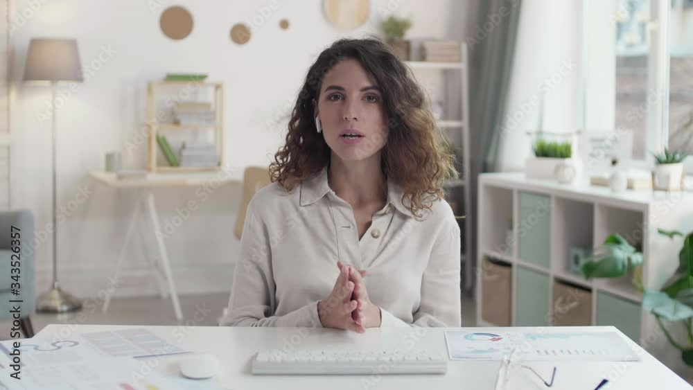 Tracking right of young pretty businesswoman wearing elegant clothes and wireless earphones sitting at desktop at computer and showing charts and graphics remotely
