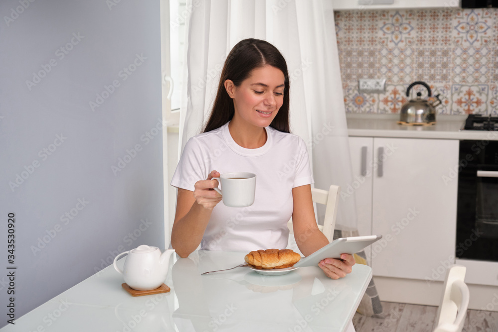 Brunette girl is drinking tea with a croissant in the kitchen , wearing a white t-shirt, white curtains in the background, laughing, watching her favourite series on the tablet, feeling great at home Brunette girl is drinking tea with a croissant in the kitchen , wearing a white t-shirt, white curtains in the background, laughing, watching her favourite series on the tablet, feeling great at home