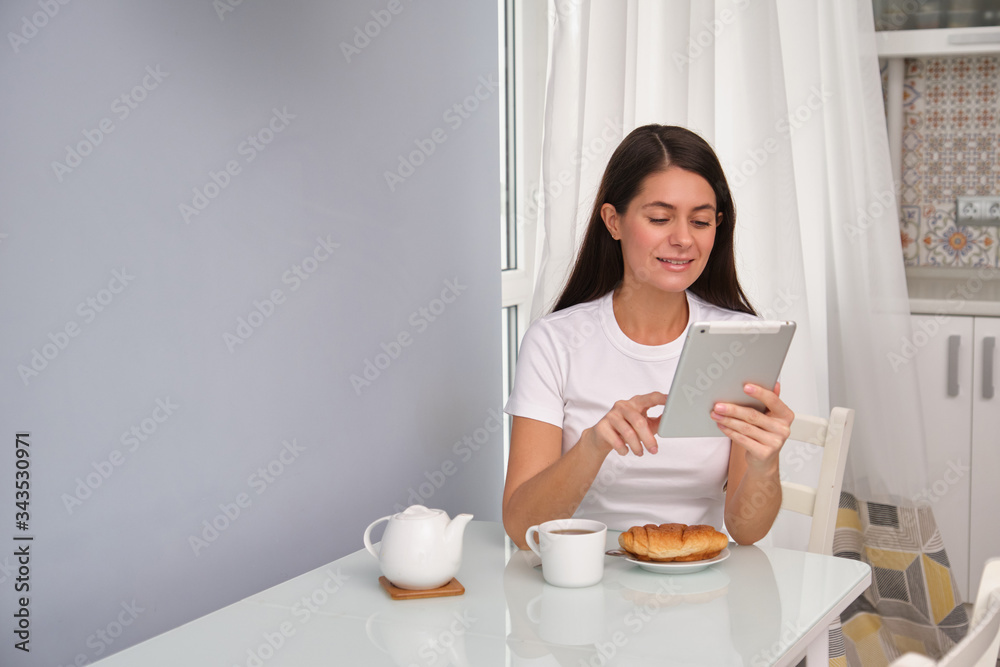 Brunette girl with long hair is drinking tea with a croissant in kitchen, wearing white t-shirt, white curtains in the background, reading an e-book on the tablet, spending good time on self-isolation Brunette girl with long hair is drinking tea with a croissant in kitchen, wearing white t-shirt, white curtains in the background, reading an e-book on the tablet, spending good time on self-isolation