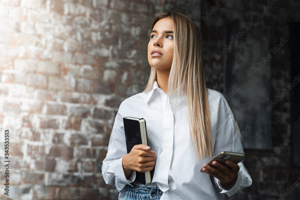 Fototapeta premium Beautiful woman standing in brick wall background. Freelance jobs for young specialists. Student with copybook