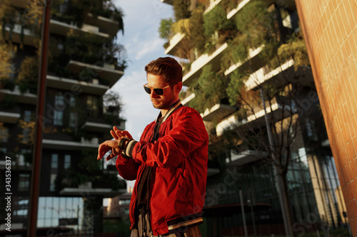 Hipster guy wear in cool clothes walking on the street looking at watch, in day time, buildings backgrounds.