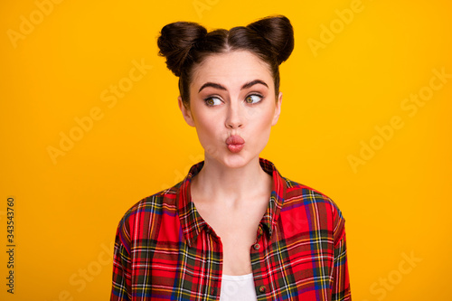 Closeup photo of pretty student lady two cute buns sending air kisses funny facial expression look shy side empty space wear casual plaid shirt isolated bright yellow color background