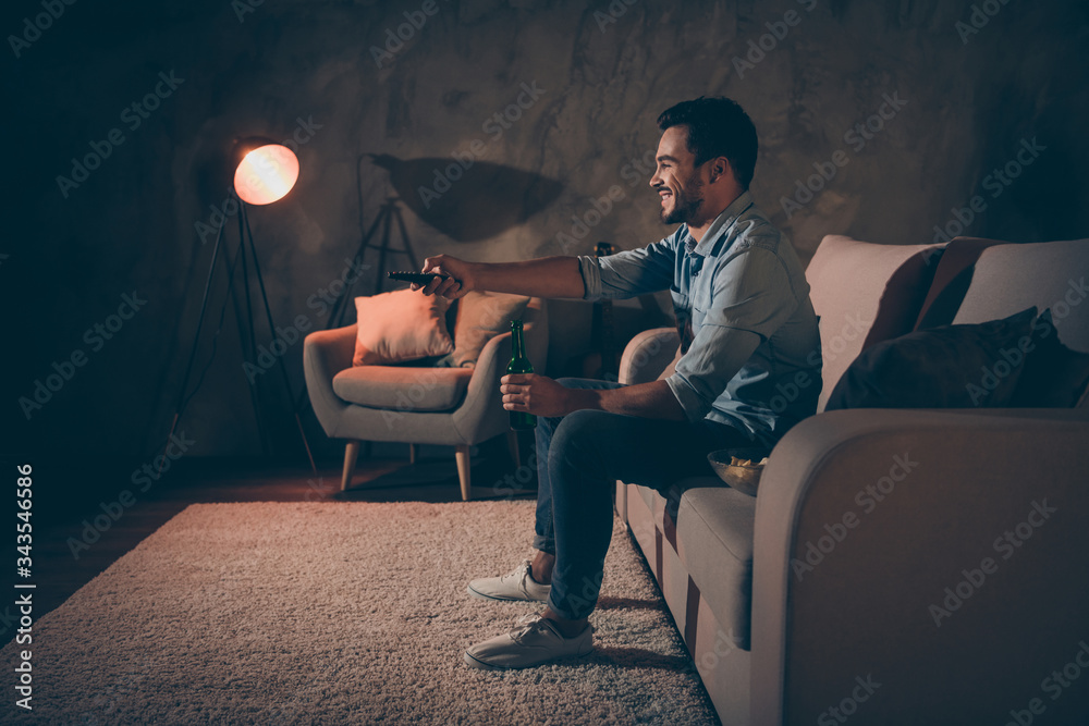 Profile side view portrait of his he nice attractive cheerful cheery brunet guy switching channel pastime drinking beverage at modern industrial loft style interior dark room indoors