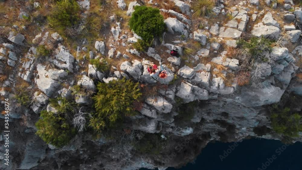 A couple of tourists in love are sitting on the edge of a cliff overlooking the sea. Edge of the world. Peak. The end of hiking trail point. Destination. and an active lifestyle. Top down, rotation A couple of tourists in love are sitting on the edge of a cliff overlooking the sea. Edge of the world. Peak. The end of hiking trail point. Destination. and an active lifestyle. Top down, rotation