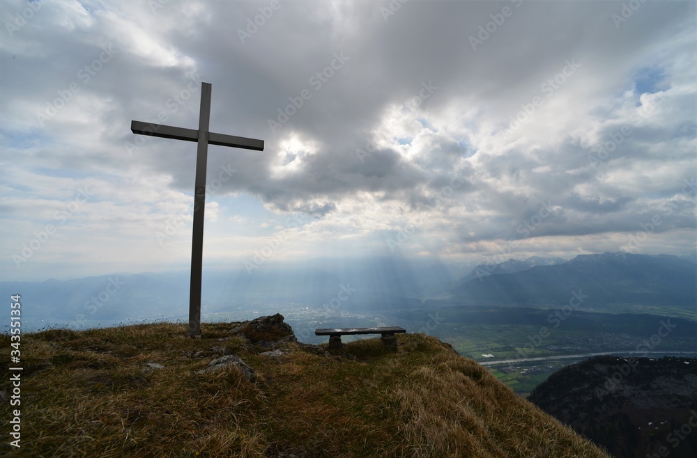 Cross on a mountain in Switzerland with a view of the valley where the river Rhine flows under a cloudy sky with sun rays