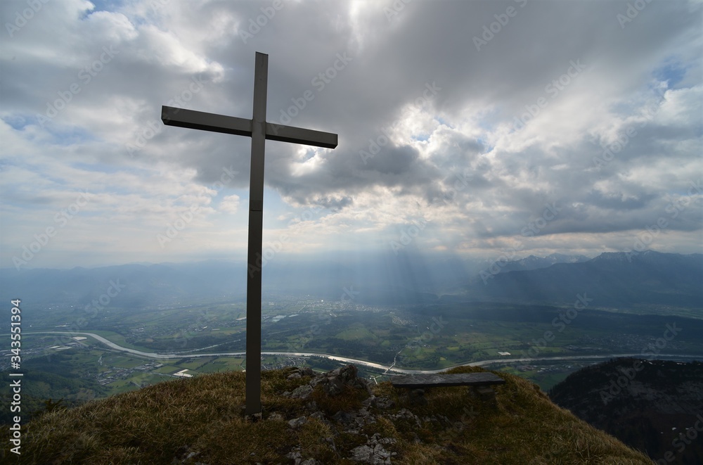 Cross on a mountain in Switzerland with a view of the valley where the river Rhine flows under a cloudy sky with sun rays Cross on a mountain in Switzerland with a view of the valley where the river Rhine flows under a cloudy sky with sun rays