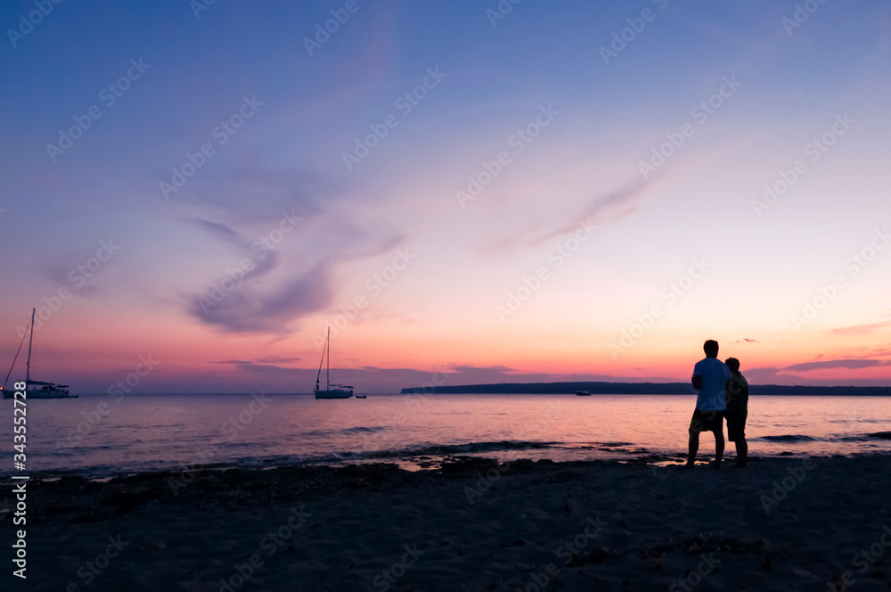 A couple watches the golden sunset in a calm sea with sailing ships in the background in Formentera, Spain, Europe