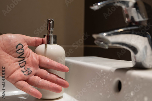 Conceptual close up shot of the left hand of a Caucasian man. On the palm there is the word covid-19, immediately next to the sink tap and the soap dispenser. Hygienize, wash away the virus.
