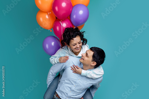 happy couple holding colorful balloons and having fun. girl is sitting on the back of boyfriend on blue background. copy space