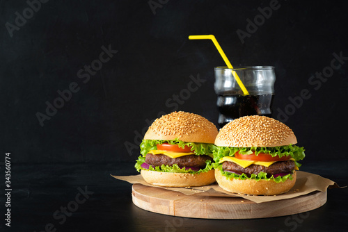 Homemade burgers, tomato ketchup and a glass of coke on little wooden cutting board over dark background.