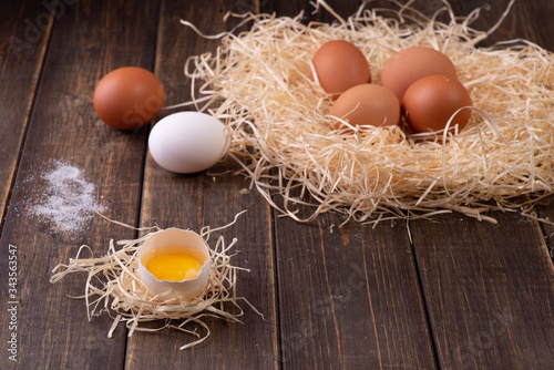 An open egg with the white and yolk on the hay, in the background in a nest of hay are whole fresh eggs, salt. Wooden background.