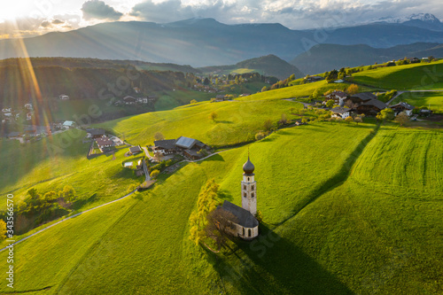 Aerial view of picturesque valley with chapel in Bolzano at sunset, Trentino, amazing green meadows of the mountains of Italy, roof tops of houses, Dolomites on background, drone flies around chapel photo