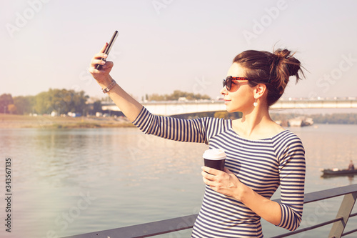 Beautiful woman using cellphone while drinking coffee near river.