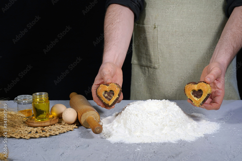 Chef with heart shaped cookies on table background with flour, eggs, oil. The concept of delicious baking Chef with heart shaped cookies on table background with flour, eggs, oil. The concept of delicious baking