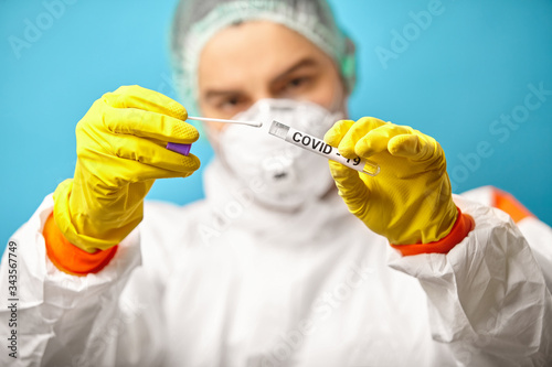 View of doctor in protective coveralls and respiratory mask, holding medical test tube with swab material taken from the mouth and nose to check for corona virus, close-up shot on a blue background.