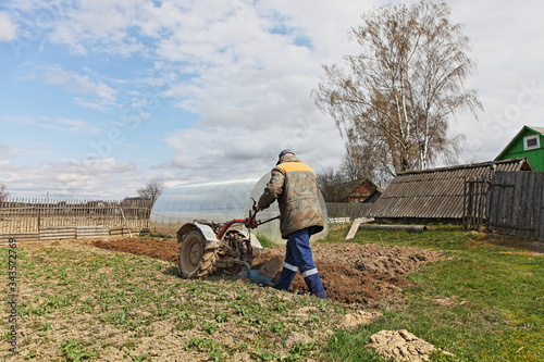 An elderly European male farmer plows a vegetable garden with a tillerblock on a spring day against the background of a greenhouse photo