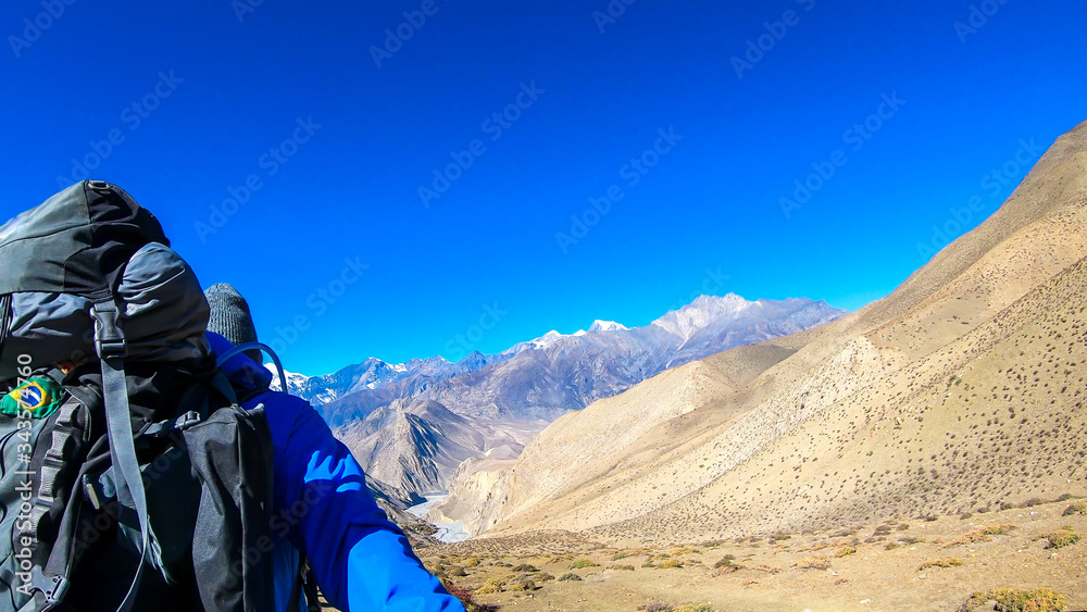 A man hiking through dry paths in Himalayan valley, located in Mustang region, Annapurna Circuit Trek in Nepal and taking a selfie. The man carries a heavy backpack. Barren slopes. Harsh landscape.
