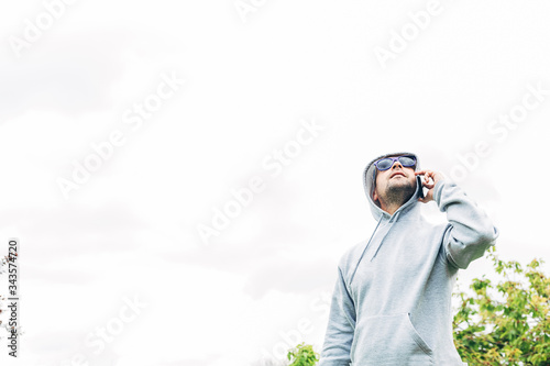 Young man in gray sweatshirt and blue sunglasses holding cell phone in his hands looking up