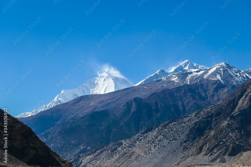 A distant view on snow capped Dhaulagiri I, seen from Mustang Valley, Annapurna Circuit Trek in Nepal. Wind blows the snow over the mountain peak. Barren and steep slopes. Harsh landscape.