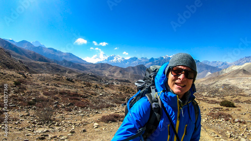 A man hiking through dry path in Himalayan valley, located in Mustang region, Annapurna Circuit Trek in Nepal and taking a selfie. The man carries a heavy backpack. Barren slopes. Harsh landscape.