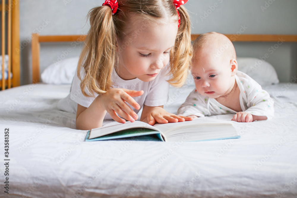 A caring older sister is reading a book to her younger newborn sister while lying on the bed. It's good to be home. A caring older sister is reading a book to her younger newborn sister while lying on the bed. It's good to be home.