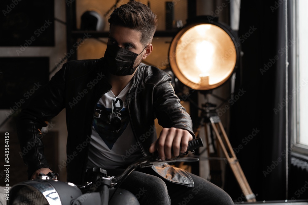Fototapeta premium Handsome brutal male biker in black mask in leather jacket sitting on motorcycle looking forward.