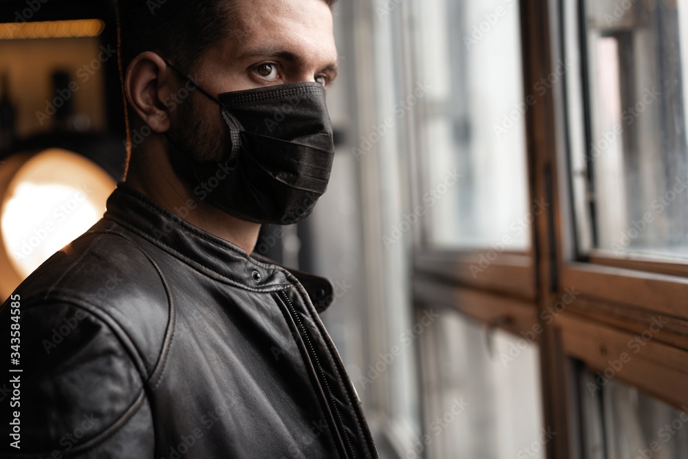 Handsome young manin black mask. Fashionable man in leather jacket standing and looking at window Handsome young manin black mask. Fashionable man in leather jacket standing and looking at window
