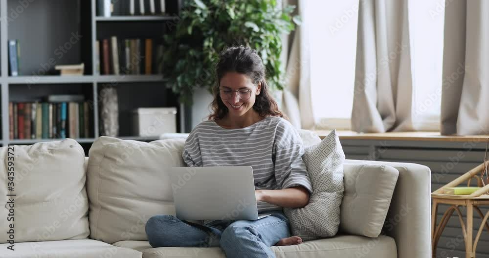 Happy young adult woman sitting in cozy living room on couch holding using laptop. Smiling casual lady chatting with friends, working or studying from home online on computer tech relaxing on sofa.