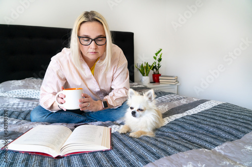 Bild auf Leinwand Blonde hair woman sitting on cozy bed with dog, holding open book and reading