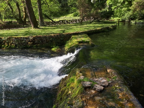 Small waterfall in the forest with green meadow and wooden fence