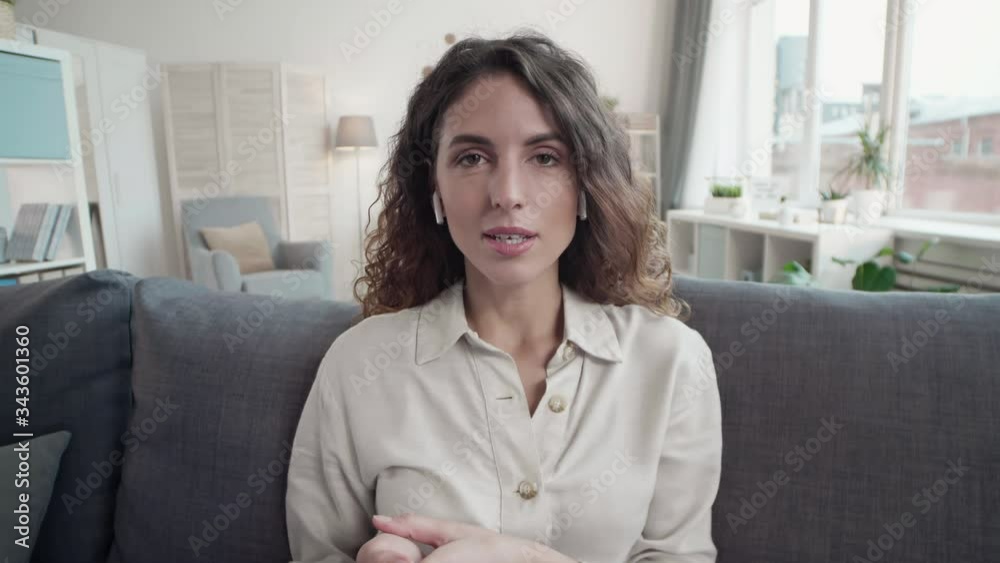 Medium shot of Caucasian young businesswoman wearing wireless earphones sitting on couch in living room and talking to business partners remotely Medium shot of Caucasian young businesswoman wearing wireless earphones sitting on couch in living room and talking to business partners remotely