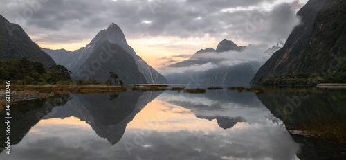 Panoramic landscape shot of sunrise in fjord with peaks shrouded in clouds and perfectly still reflection on water surface. Photo taken in Milford Sound, Fiordland National Park, New Zealand