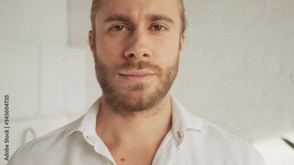 A close-up view of a calm handsome nice young man is looking to the camera in the living room at home A close-up view of a calm handsome nice young man is looking to the camera in the living room at home