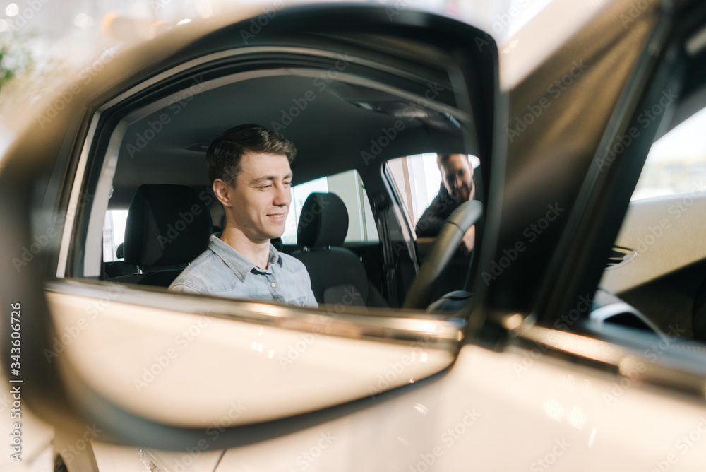 Young handsome man preparing to buy new car in auto dealership, reflection in the mirror. Professional car salesman is telling about new car model. Concept of choosing and buying new car at showroom.