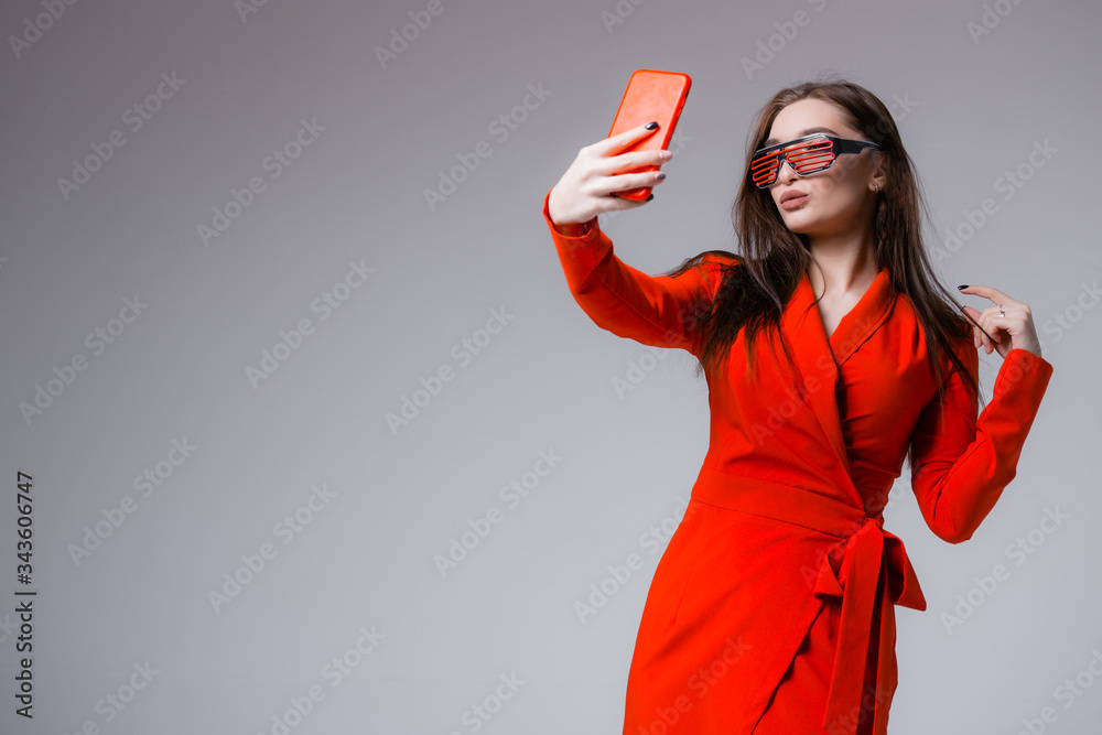 Fashionable girl with glasses in a red dress posing and taking a selfie with a new phone in her hands. Fashionable girl with glasses in a red dress posing and taking a selfie with a new phone in her hands.