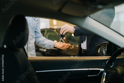 Close-up of car salesman handing over keys for new car to young man buyer in auto dealership, view from interior of car. Concept of choosing and buying new car at showroom.