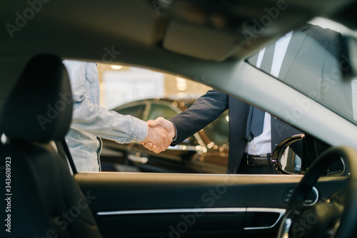 Buyer of car shaking hands with seller in auto dealership, view from interior of car. Close-up of handshake of business people. Concept of choosing and buying new car at showroom.