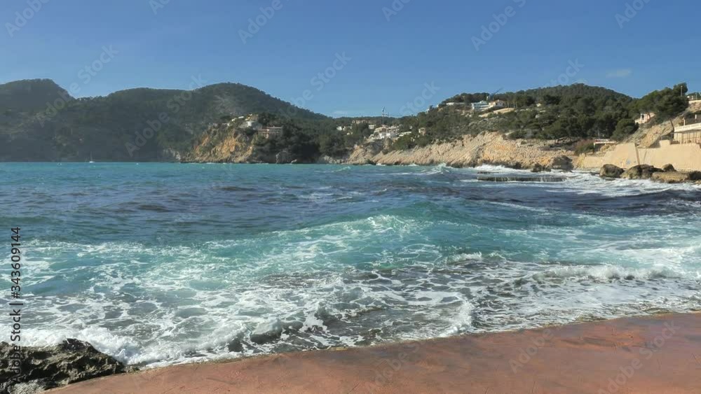 Dark hair woman walk on sea rocky bank at waving water. View of beautiful mole and beach in Camp de Mar, Majorca island, Spain Dark hair woman walk on sea rocky bank at waving water. View of beautiful mole and beach in Camp de Mar, Majorca island, Spain