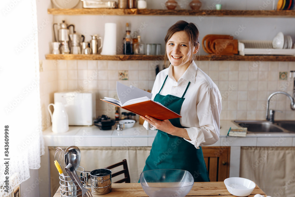Young woman standing in the middle of the kitchen with recipe book in her hands. She looking into the camera and smile. Cooking at home concept, lifestyle. Young woman standing in the middle of the kitchen with recipe book in her hands. She looking into the camera and smile. Cooking at home concept, lifestyle.
