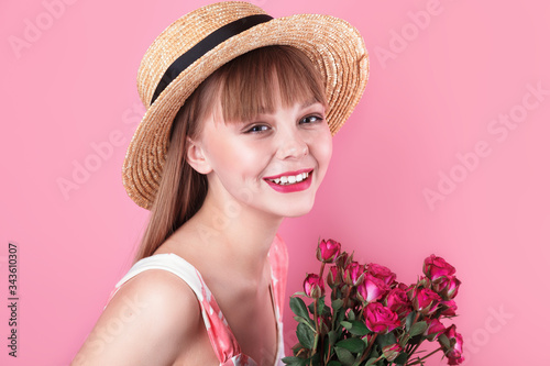 Smiling young woman in summer dress and straw hat holding bouquet of roses on pink background
