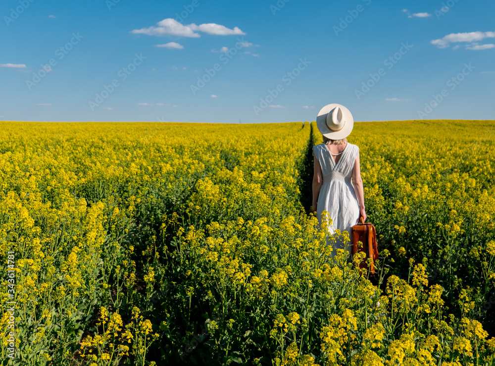 Beautiful woman in dress with suitcase in rapeseed field in spring time