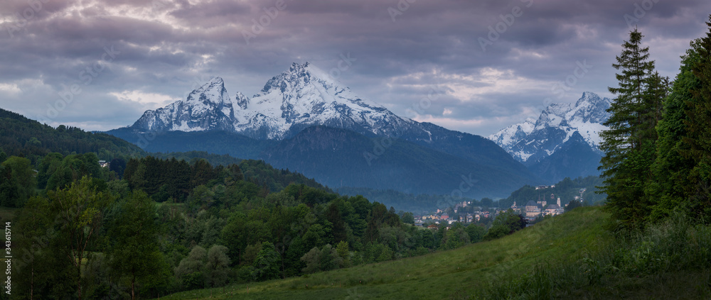 Snow covered mountain peak panorama of Watzmann and city Berchtesgaden with clouds during sunrise, green forest and fields in foreground, Bavaria.