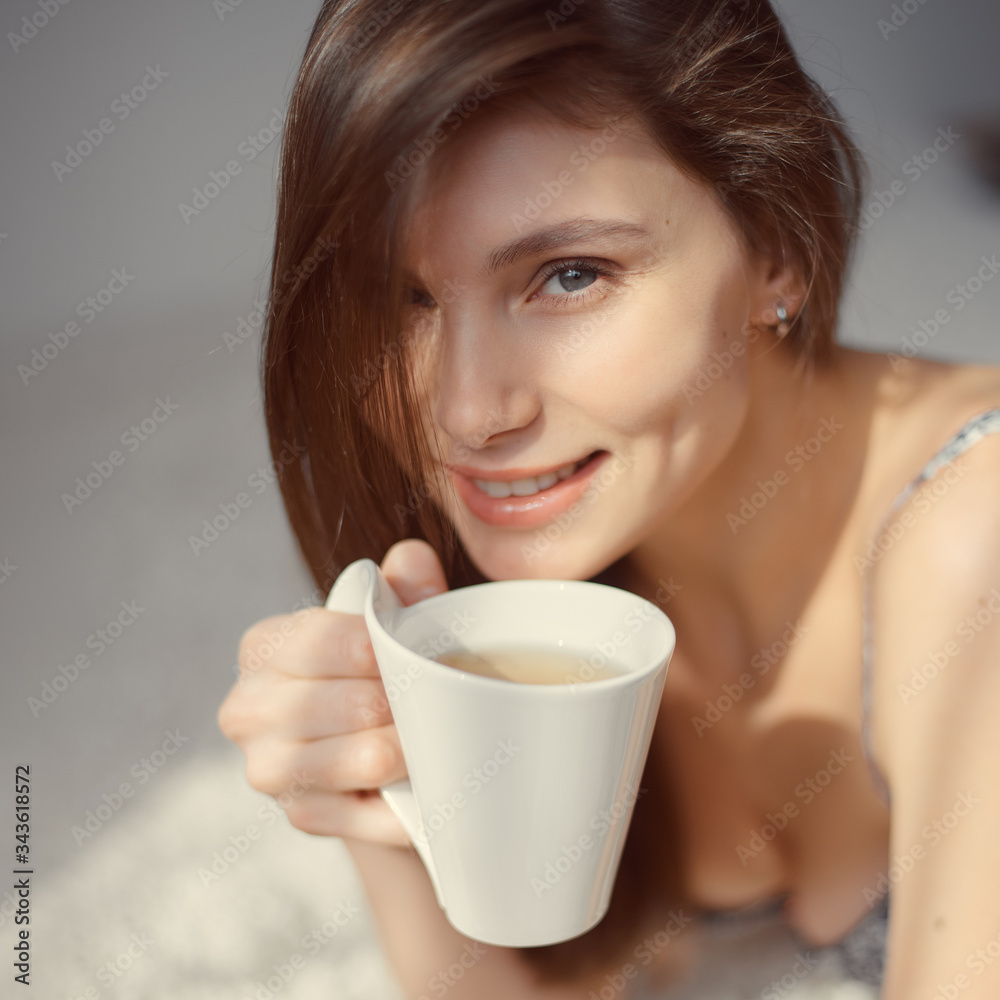 young woman with a cup of tea in her hands looks at the camera. the room is flooded with sunlight young woman with a cup of tea in her hands looks at the camera. the room is flooded with sunlight