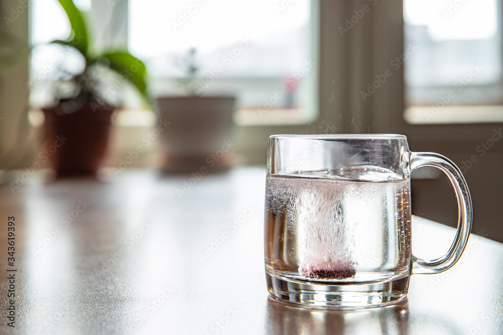 Sparkling tablet throw into a glass transparent glass, the glass is on a table, a pill for cold of claret color, bubbles rise up, splashes take off from a glass, flowers is on background, foam