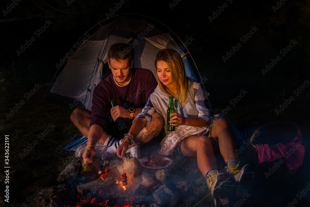 Young couple baking sausages on the campfire and drinking beer in the forest hill in the dusk Young couple baking sausages on the campfire and drinking beer in the forest hill in the dusk