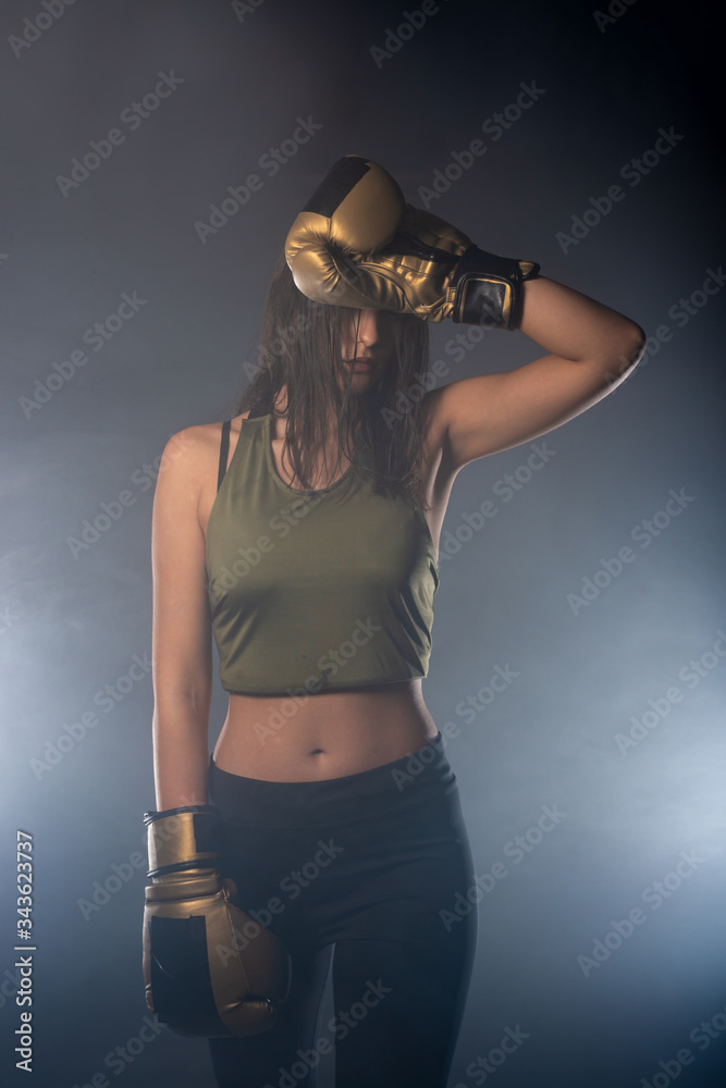 Smokey portrait of an attractive fit girl wearing boxing gloves posing with her arm over her head.