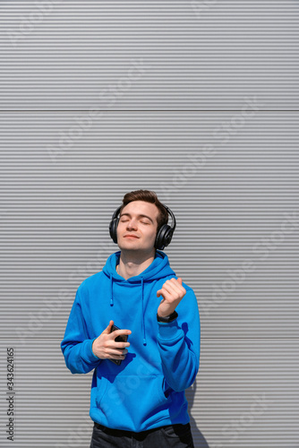 happy young student with smile and closed eyes, in headphones dancing with smartphone in his right hand against a gray wall in the afternoon, wearing blue hoodie, black jeans and black fitness tracker